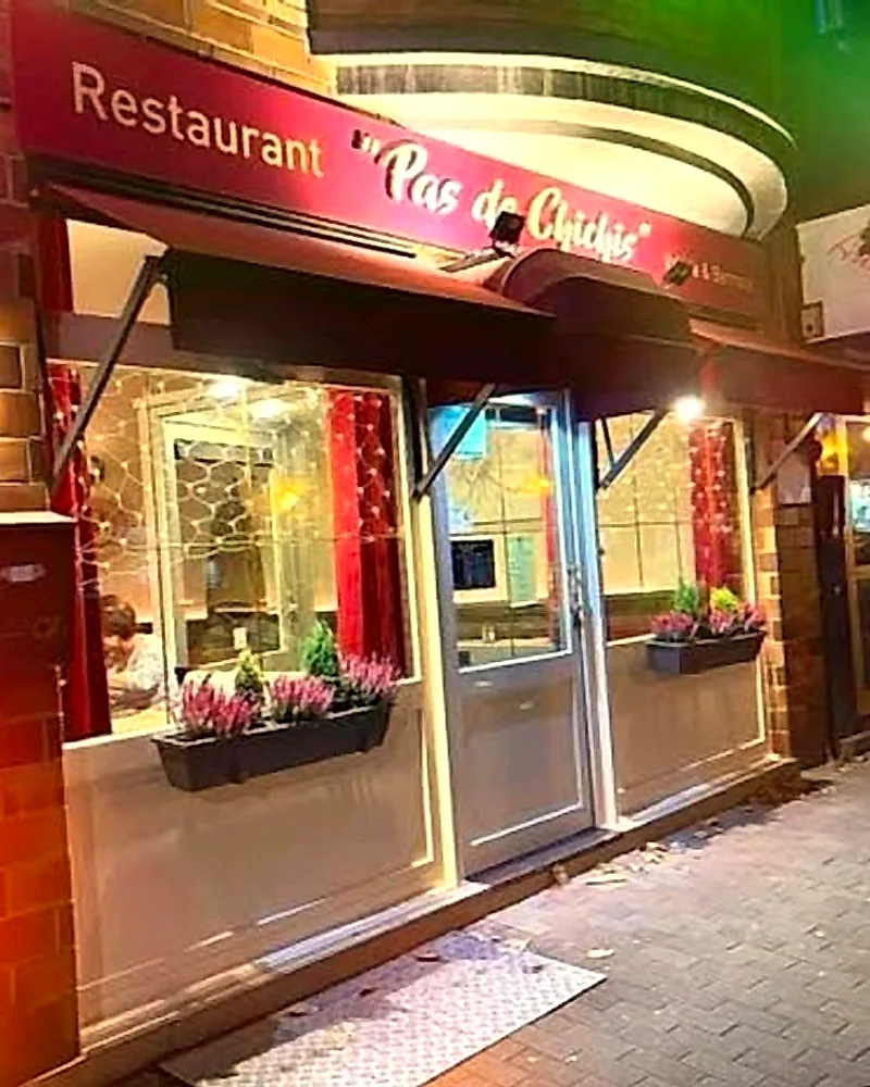 Façade du restaurant Pas de Chichis, le soir : enseigne « Restaurant Pas de Chichis », auvents bordeaux, jardinières de bruyère et porte vitrée